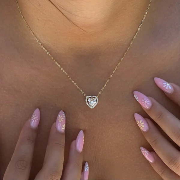 תמונת קלוז-אפ של שרשרת לב כסף משובצת אשלי בציפוי זהב עדינה, משובצת אבנים נוצצות, על צווארה החשוף של אישה. ידיה שלובות בחזית, וציפורניה הצבועות בלק ורוד מעוטרות בפרחים לבנים וצהובים קטנים.
