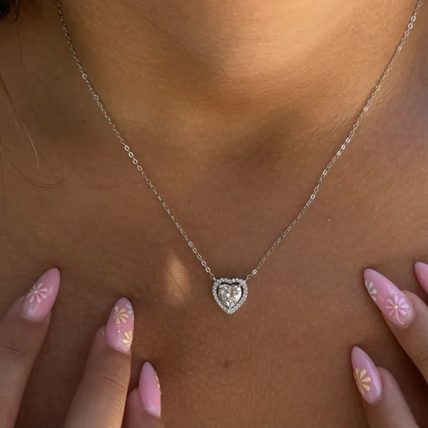 תמונת קלוז-אפ של שרשרת לב כסף עדינה, משובצת אבנים נוצצות, על צווארה החשוף של אישה. ידיה שלובות בחזית, וציפורניה הצבועות בלק ורוד מעוטרות בפרחים לבנים וצהובים קטנים.